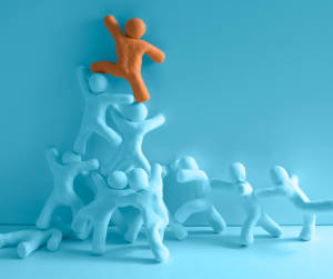 An orange clay figure triumphs at the peak, supported by a pyramid of numerous blue clay figures, symbolizing teamwork, leadership, and collective success.
